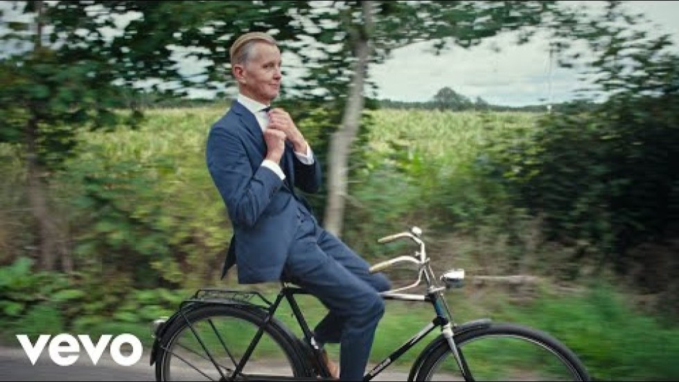 retro-style footage of young children pantomiming grown-up activites like reading the newspaper and driving a car, while an adult man rides a bicycle down a country road