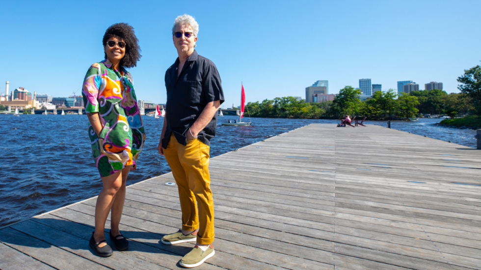 a woman in a colorful dress and a man in a blue shirt and gold trousers stand on a dock in the Charles River.