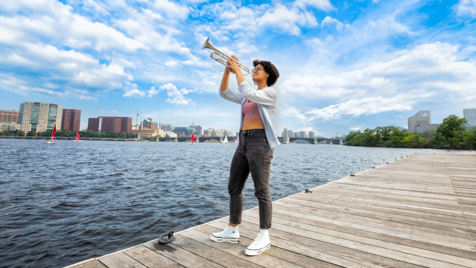 a young multiracial woman plays her trumpet on a dock in the Charles River. She wears glasses, a cropped peach shirt and gray jeans, and thick-soled white sneakers. 