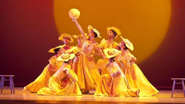 A group of dancers from Alvin Ailey American Dance Theater perform on stage. They all wear yellow dresses, yellow hats, and are congregated around one dancer holding a yellow fan aloft.