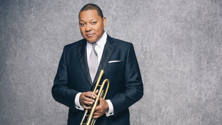 Wynton Marsalis, a black man wearing a dark suit with a white shirt and light grey tie, stands in front of a grey background while holding his trumpet