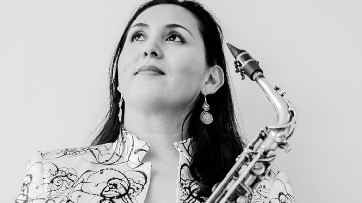 A woman, holding a tenor saxophone, looks up and out of frame.