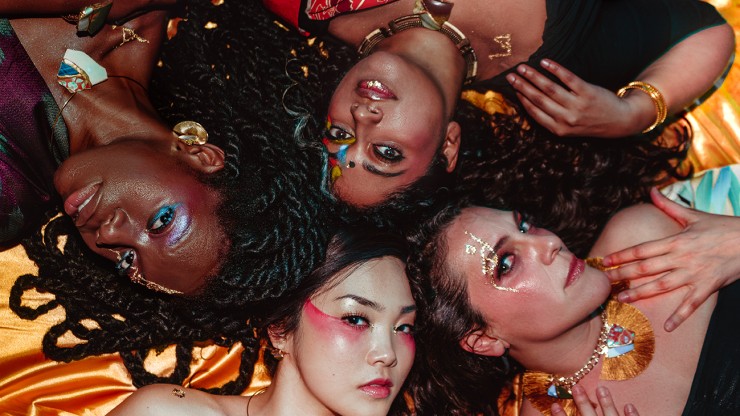 four women lie on the ground, their heads together. They are dressed in colorful international clothing and wear dramatic makeup
