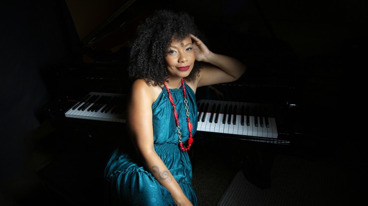 an afro-cuban woman in an elegant dark turquoise dress leans an elbow over the keys of a piano, against a black background in a dark studio