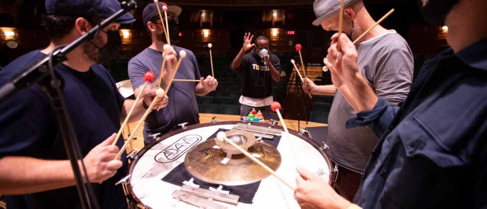 gathered around a bass drum turned on its side with percussion instruments on top, four members of a percussion duo and a beatboxer rehearse