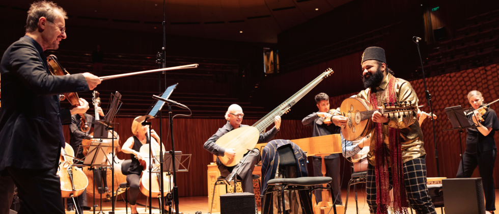 white man in a black suit conducts a chamber orchestra, featuring a smiling Egyptian Australian man with a curly mustache, a thick beard, and a cylindrical maroon hat