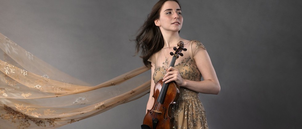 Before a gray photo backdrop, a young Armenian woman in a tan dress holds a violin. An embroidered scarf billows behind her.