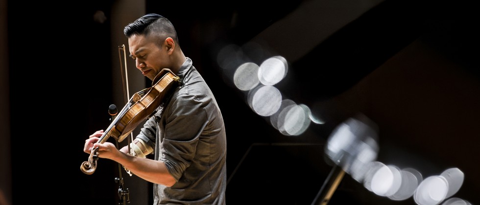 A man with dark hair performs violin with a visible limb difference on his left side next to a microphone. 