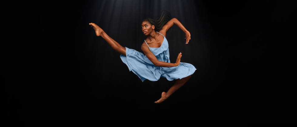 a dark skilled black woman leaps beofre a black background, one leg extended, one tucked under. Her arms, swept to her side, make a circle.