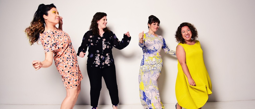 Aizuri Quartet, four women in patterned and solid clothing, stand in front of a white wall dancing and smiling. 