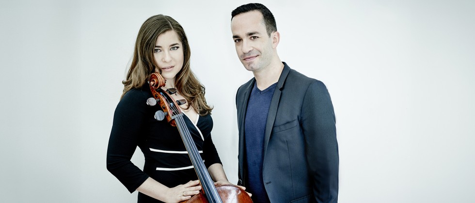 A man in a dark suit and woman with long brown hair holds her cello as they stand close together in front of a white background. 
