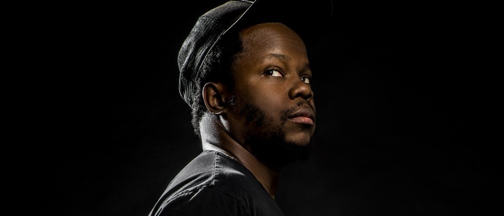 Black man, Ambrose Akinmusire in a black shirt and hat in front of a black background. 