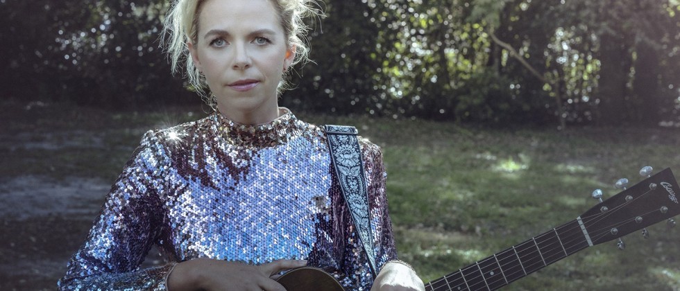 A white woman with blond hair and a metallic sequins dress, and an acoustic guitar stands in front of a tree-lined field.