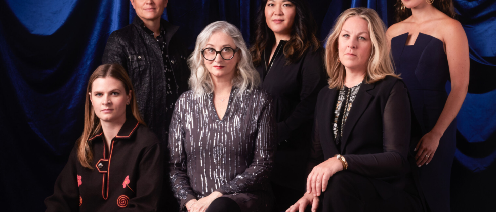 Six women, standing and seated in front of a blue backdrop
