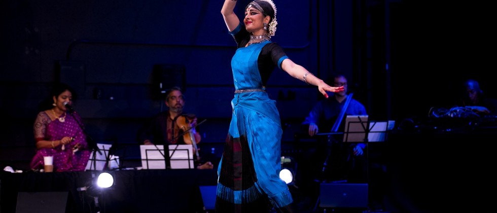 a female dancer of Indian heritage, dressed in an ornate blue traditional outfit and adorned with gold jewelry, steps forward, one arm raised with her thumb and first finger pinched together