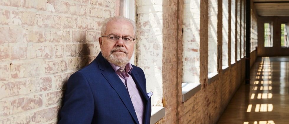 An older, white man in a navy jacket leans against a light brick wall lined with windows. 