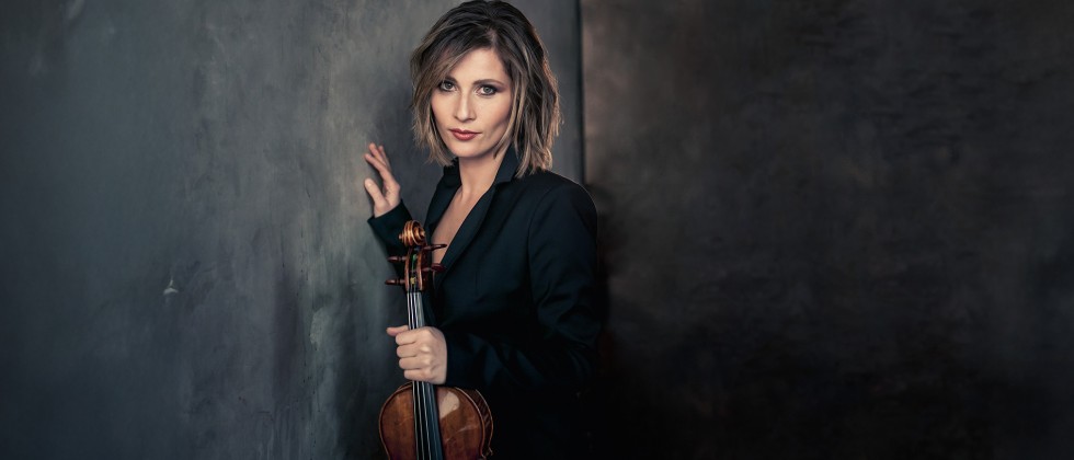 A white woman with shoulder length brown hair leans against a gray painted wall holding her violin while wearing a dark suit jacket. 