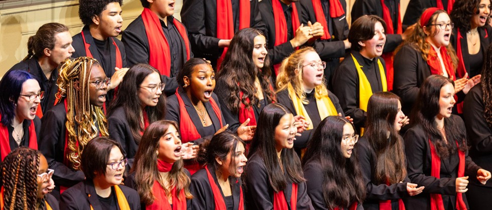 On a series of risers, a large multicultural chorus of young people stand singing, holding binders, and wearing colorful red, orange, and yellow stoles. 
