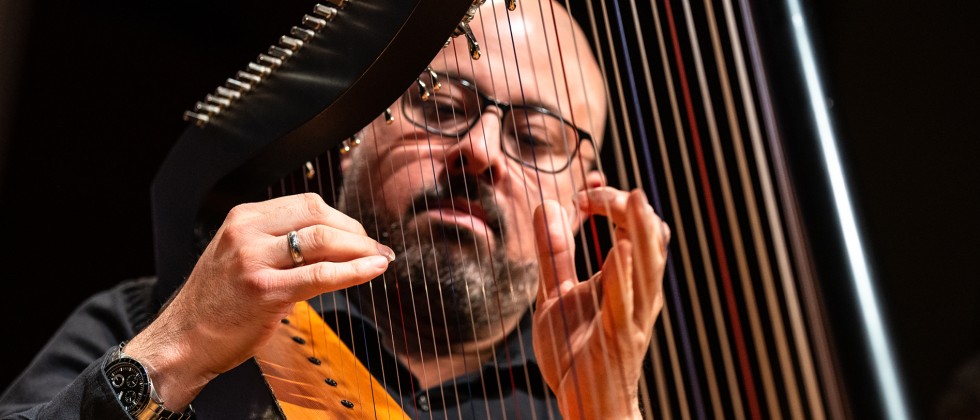 A Venezuelan-born man with facial hair and glasses can be seen playing the harp through the strings of the instrument. 