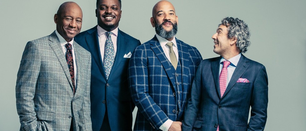 four men in suits smile warmly, the rightmost turned to the side to look at the group.