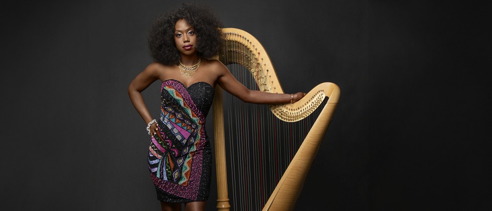 A Black woman poses with a golden harp in front of a dark background in a short, strapless dress with a geometric pattern. 