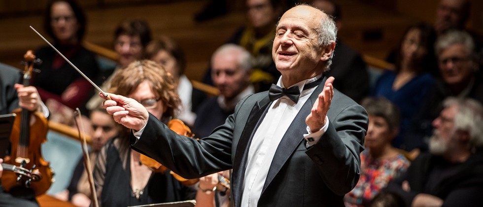 An older white man in a suit and a bow tie conducts an orchestra with arms outstretched in front of a crowd. 