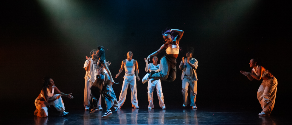 some standing, one crouched, dancers in streetwear gather behind a woman who jumps, one hand to her head and her legs bending behind her
