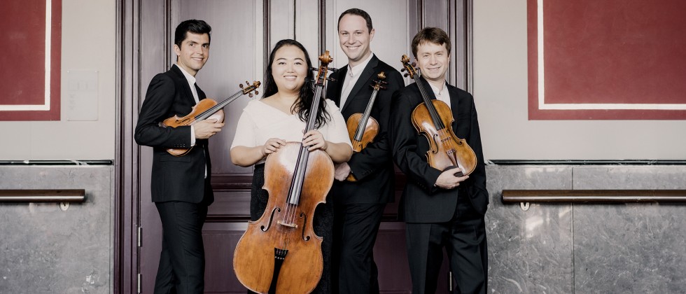 A string quartet of 3 women and a man stand in front of closed light door. 