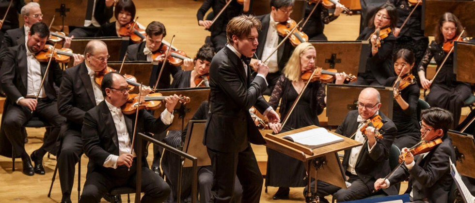 A young man conducts a large orchestra on a red podium; behind him the violin section is visible in formal concert attire. 