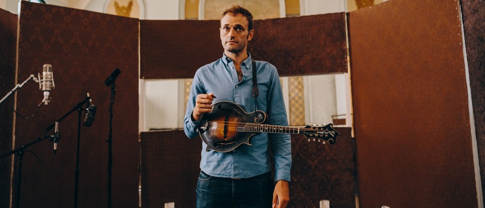 a mandolin player, in a recording booth within a historic church, pauses with his pick in hand and his instrument across his body