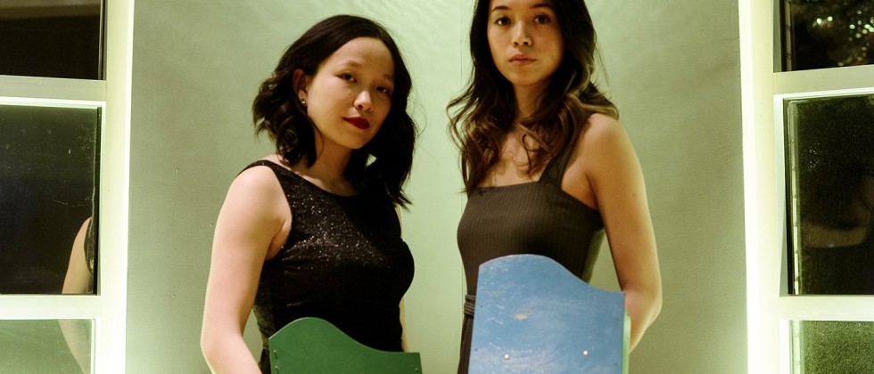 two young asian-american women in sleeveless dresses hold toy pianos as they stand under a small disco ball. 