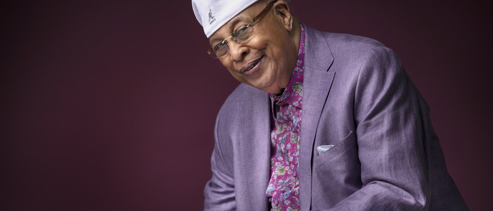 an older cuban man in a purple jacket and a flat cap smiles, with his hands poised over a keyboard