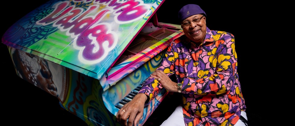 An older black man in a colorful purple, pink, and yellow top and a purple cap leans on a grand piano covered in colorful graffiti. 