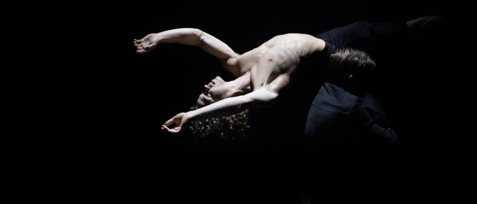 a circus performer, lifted by a barely-visible colleague, arcs his torso backward, his pale skin luminescent under a single spot of stark white lighting