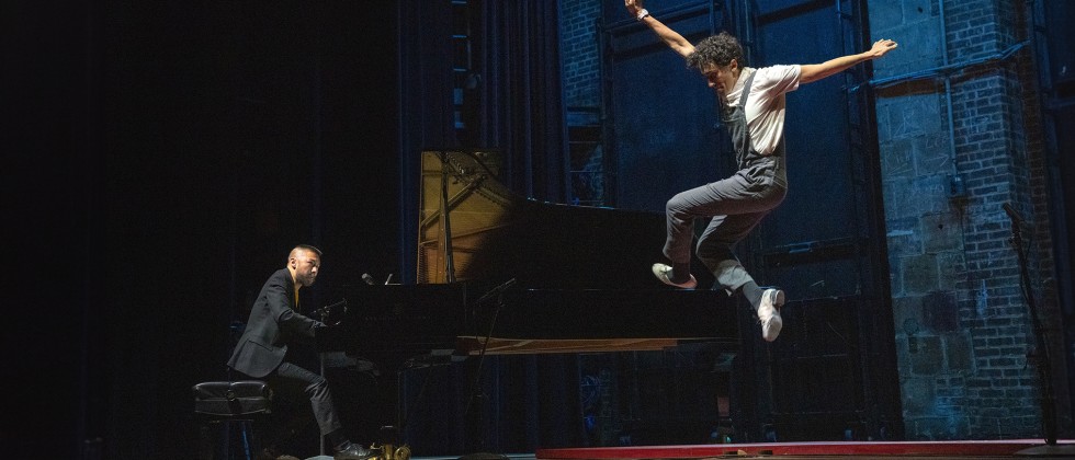 Two performers are pictured on a stage; one sits at a grand piano while the other is pictured high in the air, arms outstretched. 