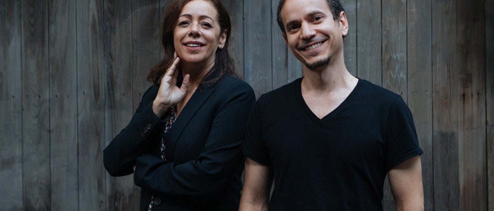 A brazilian woman, her hand to her jaw, and a cuban man wearing a t-shirt with his hands in the pockets of his jeans, in front of a concrete wall. They both look amused, as if the photographer said something funny.