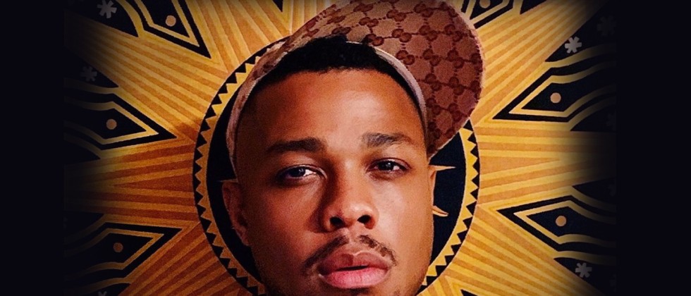 Davone Tines, a black man looks at the camera standing in front of an orange and black banner. 