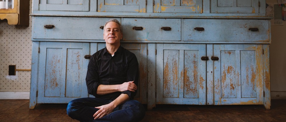 A white man in dark jeans and a black button-down shirt with the sleeves rolled up leans back against a rustic cupboard with flaking blue paint