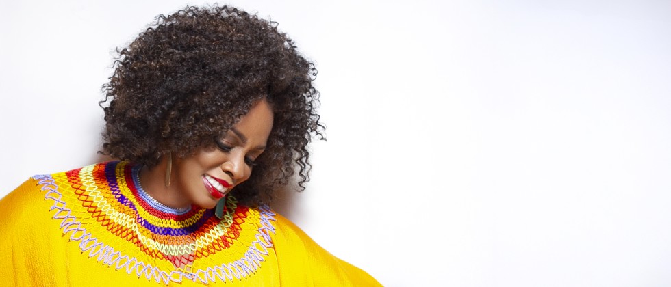 A black woman with shoulder length hair wears gold earrings and a bright yellow shirt with a colorful collar while looking downward. 