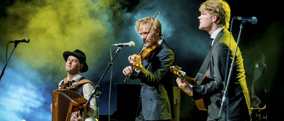 Three musicians stand diagonally, playing a guitar, violin, and accordion respectively in front of a greenish-blue fog. 