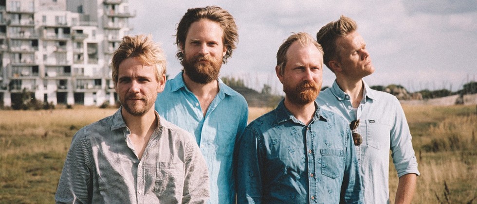 four blond men in casual shirts stand outdoors in a field in the sun. the sky behind them is gray, and there is an apartment building in the far background.