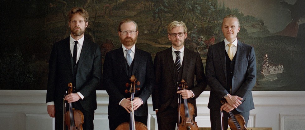 Four white men in a string quartet stand in a row in dark suits holding their instruments in front of several nature murals. 