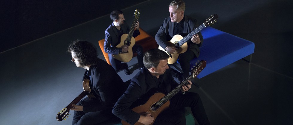 Four Irish guitar players sit spread out between three ottomans, facing different directions while wearing concert black. 
