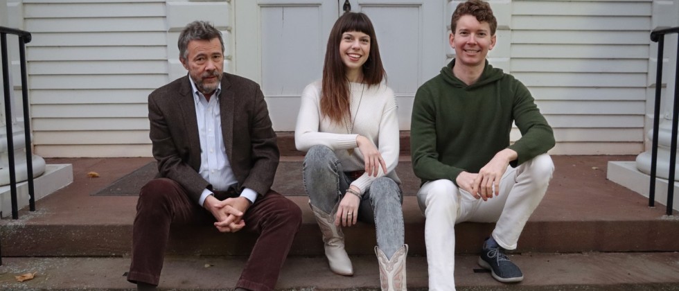 A trio sits in front of a white building. The woman smiles wearing white cowboy boots; a man wears a brown suit; and a man smiles broadly. 