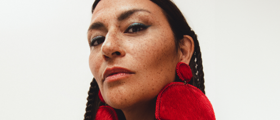 An Indigenous woman, in a 3/4 profile, looks down toward the camera. She wears red semicircular earrings made of fur with a beaded fringe.