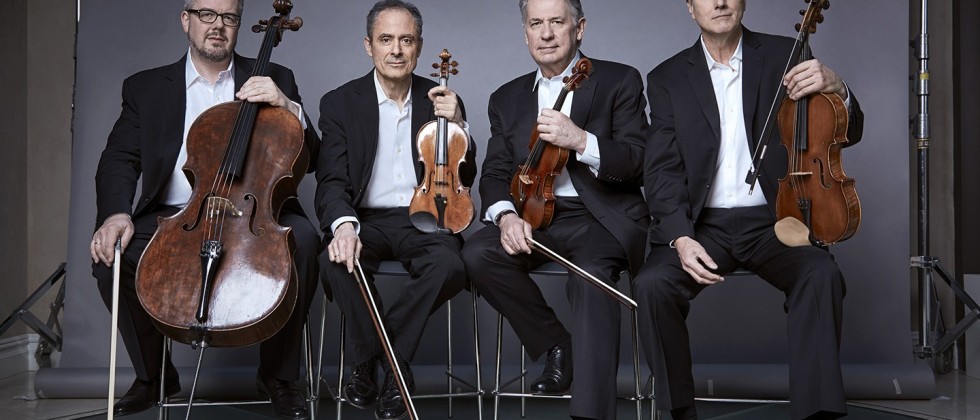 four gray-haired white men in matching gray suits hold their stringed instruments before a gray photo backdrop