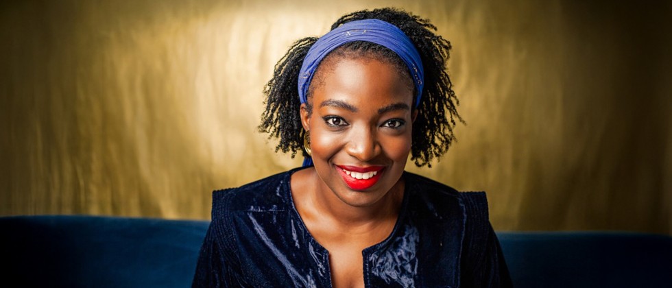 A smiling black woman with a blue headband, red lipstick, gold earrings, and a velvet-like dark blue jacket. 