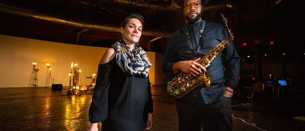 A white woman holding percussion mallets and a black man holding a saxophone stand in a studio dimly lit with golden light