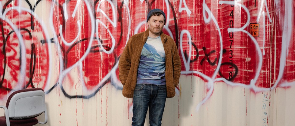 A white man stands in blue jeans, a t-shirt with mountains, and a brown fuzzy jacket in front of a wall of graffiti. 