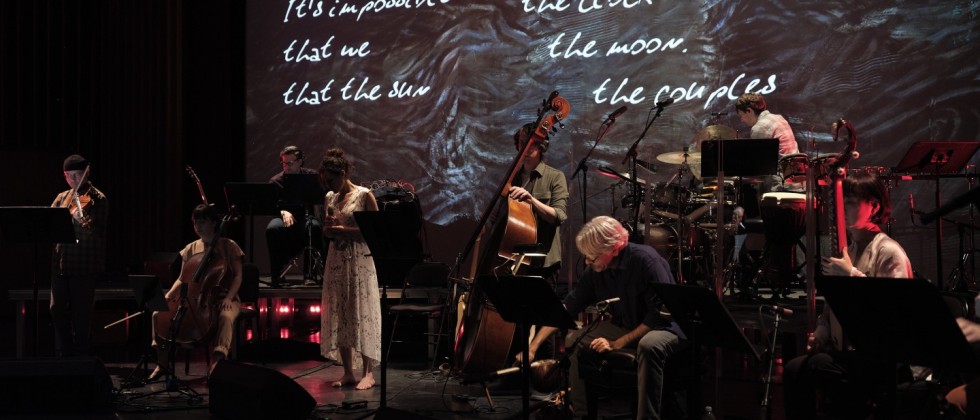 The cast of Falling Out of Time, a group of thirteen men and women standing outside on a stone path with orange trees behind them, with an inset photo of Osvaldo Golijov in the bottom right corner. 